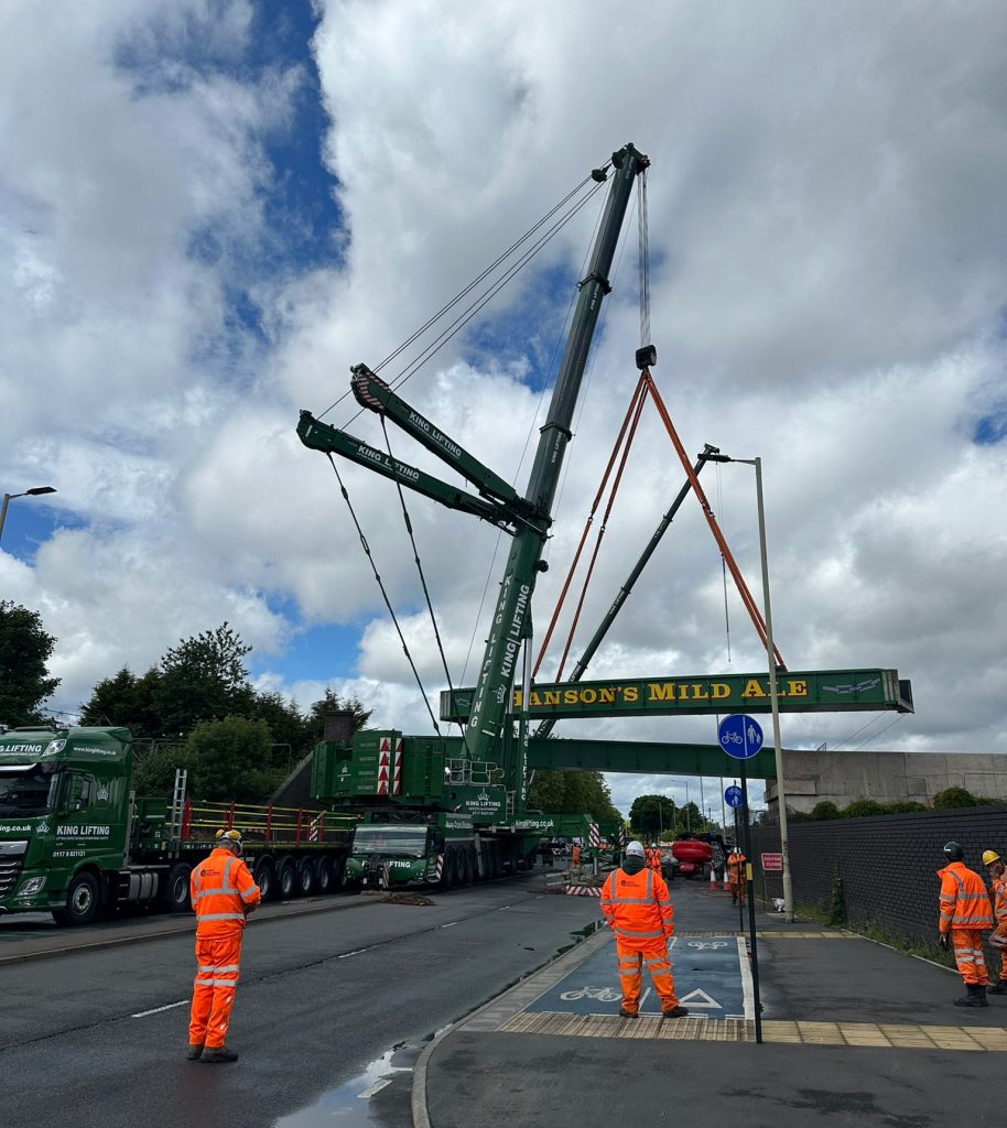 King Lifting crane team installing a Hanson's Mild Ale sign bridge with large green crane on urban street
